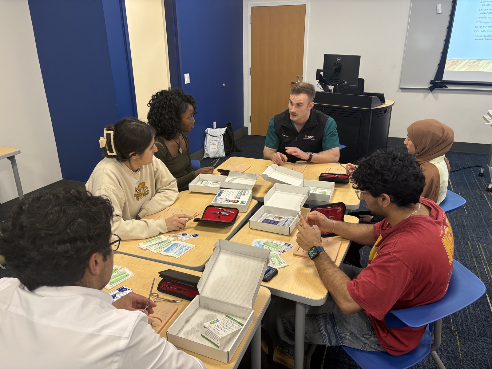 A MedRise mentor demonstrating suturing technique to students seated around a table at the University of Memphis